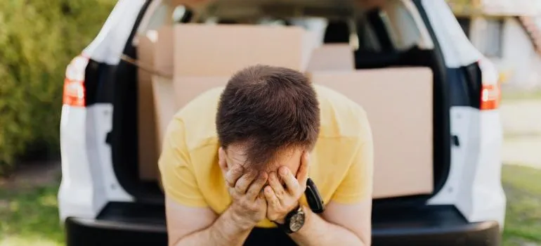 Man with his face in his hands, and a car packed with boxes behind him.