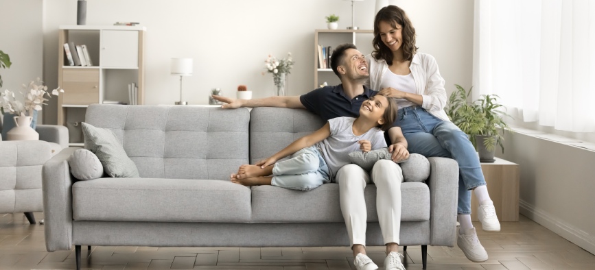Happy couple of young parents and little daughter kid sitting and relaxing on sofa, resting on comfortable couch in modern cozy home interior, talking, laughing, smiling. Full length wide shot